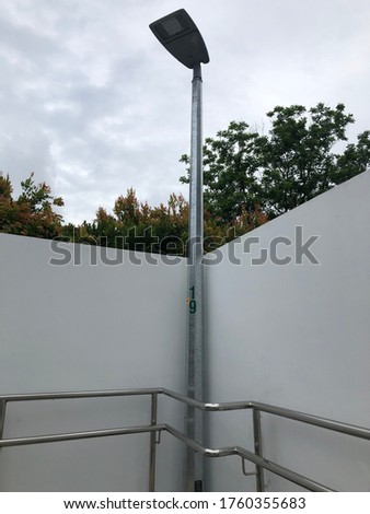 Lamp post in front of a wall barricade and behind silver railings metal with green trees in the background and white clouds in the background