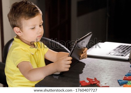 3 year old boy with yellow polo plays with a tablet sitting in the kitchen table