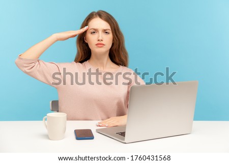 Yes sir! Obedient responsible young woman employee sitting at workplace with laptop and giving salute, listening to boss order, corporate discipline. indoor studio shot isolated on blue background Royalty-Free Stock Photo #1760431568