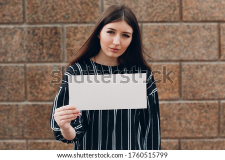 Young woman holds white paper in hand stone background. Girl with white blank template sheet with empty space