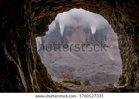 Dolomites Alps. Tre Cime di Lavaredo. Italy. Three dolomite grey peaks of ancient mountains stands on the alpine desert wrapped by white clouds. Horizontal view from the cave