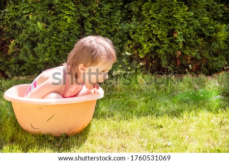 A little kid bathes in an orange basin in the summer in hot weather against a background of green bushes and grass. The girl sits sideways, stuck out her tongue