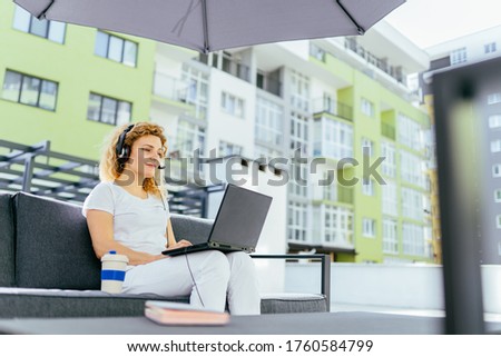 Serious young woman in headphones, wearing white casual outfit checking schedule in organizer while working remotely with laptop in terrace of coffee shop.