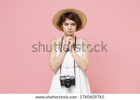 Pleading young tourist woman in summer dress hat with photo camera isolated on pink background studio. Traveling to travel weekends getaway. Air flight journey concept. Holding hands folded in prayer