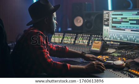 Energetic Male Audio Engineer / Producer Working in Music Recording Studio, Mixing Tracks on Control Desk and Software to Create Hit Song Track. Artist Musician Enter His Studio at Sits at Workdesk Royalty-Free Stock Photo #1760613896