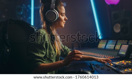 Portrait of Beautiful Female Artist Musician in Music Recording Studio, Uses Headphones.  Successful Female Audio Engineer Uses Mixing Board Create Modern Song. Royalty-Free Stock Photo #1760614013