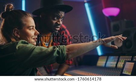 Stylish Producer and Audio Engineer Working together in Music Recording Studio on New Album, Talk, Use Control Desk Equalizer, Mixing Board and Software to Create Hit Song. Artist and Musician Collab Royalty-Free Stock Photo #1760614178
