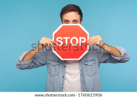 Way prohibited! Portrait of man in worker denim shirt peeking out of Stop road traffic sign, showing symbol of restrictions and forbidden access. indoor studio shot isolated on blue background