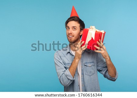Portrait of curious man with funny party hat holding gift box near ear and listening, guessing present, in anticipation of interesting birthday surprise. indoor studio shot isolated on blue background
