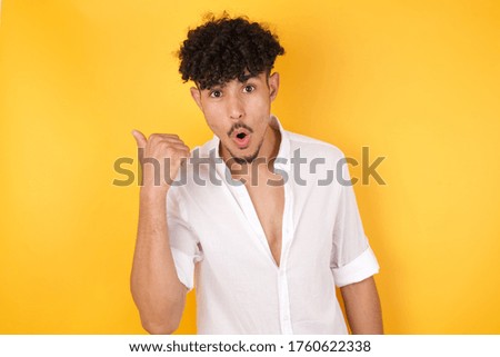 Horizontal shot of pretty young joyful shocked male points with thumb away, indicates something on blank wall isolated over yellow background. Check this out.  