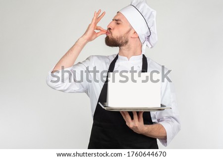 The gesture means delicious. The chef holds a tray with an empty sign. Space for text. The chef makes an approving gesture with his hand. The taste of the product is highly appreciated.