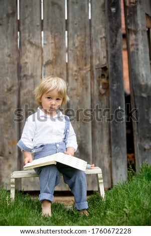 Blond toddler boy, reading book in garden in front of old wooden gate