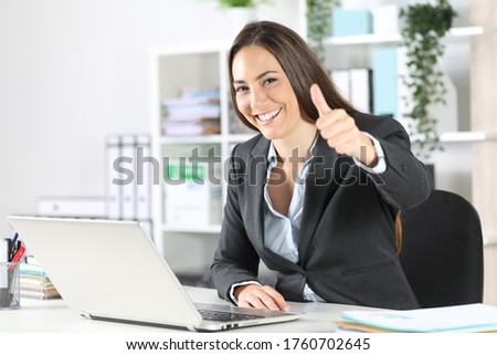 Happy executive woman with thumbs up looking at camera sitting on a desk at the office
