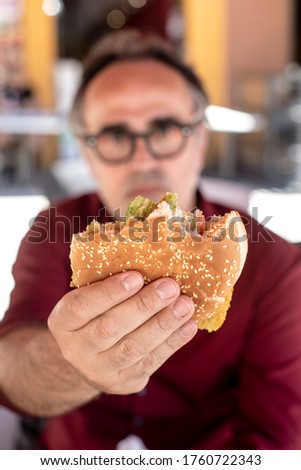 Man with black hair and eyeglasses shows in the foreground the sandwich with the freshly bitten cutlet