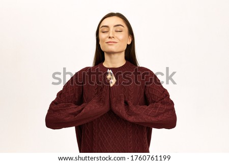 Portrait of charming young woman with dark long hair posing in jumper gesturing, pressing palms together, keeping eyes closed, having calm peaceful facial expression, praying, being grateful