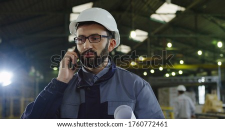 Caucasian factory worker in a helmet talking on the smart phone in the middle of big industrial room. Indoor. Portrait. Close up