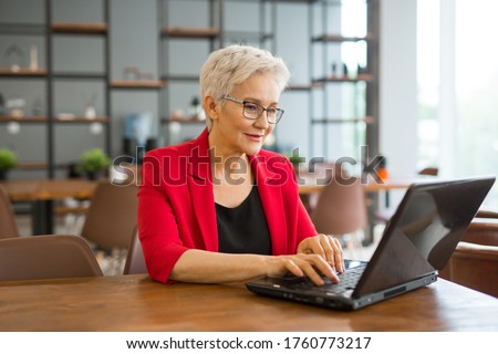 beautiful aged woman in a red jacket works in the office with a laptop Royalty-Free Stock Photo #1760773217