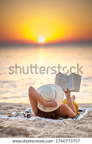 Young woman reading a book at the beach while the sun rise. She's wearing a orange dress and white hat Royalty-Free Stock Photo #1760773757