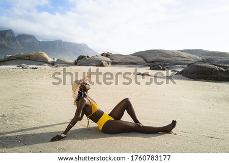 Side view of female in bikini with sun hat sitting at the beach. African woman resting on the beach on a summer day.
