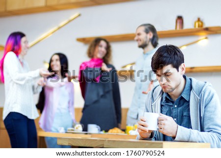 Closeup portrait of sad man, outcast is sitting alone at table in cafe. Teenagers, students are mocking, scoffing, pointing at guy. Influence of society on mental health. Bullying concept.  Royalty-Free Stock Photo #1760790524