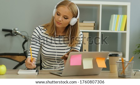 Young beautiful woman listening music in headphones and making some notes in the note book in the modern office.