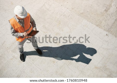 An engineer in an orange vest and a white construction control helmet conducts an inspection with a tablet in his hands, top view