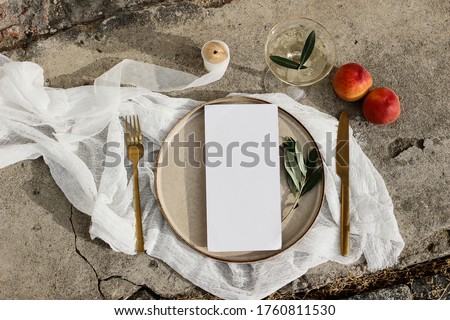 Festive wedding summer table setting. Golden cutlery, olive branch, glass of wine, peaches fruit and porcelain dinner plate on grunge concrete background. Blank menu card mockup. Flat lay, top view.