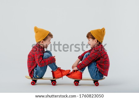 Side view of cheerful twin brothers wearing similar shirts and jeans relaxing on skateboards on white background and looking at each other Royalty-Free Stock Photo #1760823050