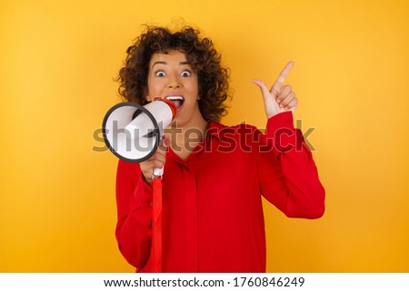 Young arab woman with curly hair wearing red shirt holding a megaphone  over yellow background. holding finger up having idea and posing 