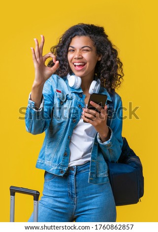 Vertical shot of happy cute casually dressed tourist girl with backpack and mobile phone winking and showing ok gesture. Isolated over yellow background.