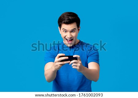 Excited handsome young male gamer playing online game on smartphone isolated on blue background