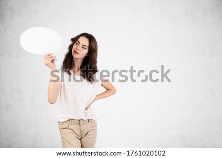 Half length portrait of young dark haired woman in casual clothes holding speech bubble near concrete wall. Concept of communication and advertising. Mock up
