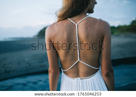 Back view of anonymous slim female in white beach dress standing on wet sandy coast near waving sea while enjoying holidays on Bali and looking away