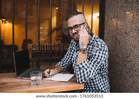 Positive young male blogger in eyeglasses and checkered shirt having phone conversation and writing information in planner while sitting at wooden table with laptop in outdoor cafeteria
