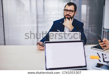 Thoughtful male employee in spectacles pondering on financial research during collaborative working process at desk with mockup laptop computer, blank netbook with copy space for business website