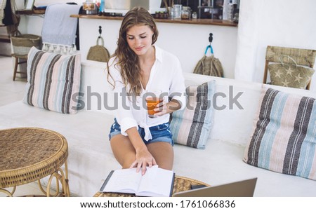 Pensive tanned female in casual wear reading book and enjoying juice while relaxing on cozy couch in vintage restaurant in summertime
