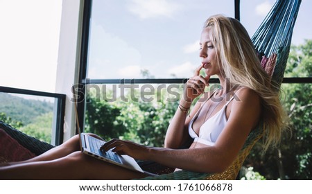 Side view of tranquil tanned female in white bikini chilling in comfortable hammock and using laptop on terrace at resort