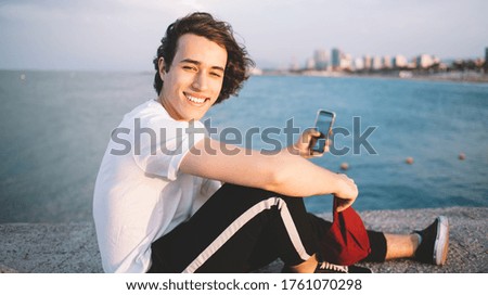 Side view of young guy with cap in hand on background of clear water and cloudy sky using phone and taking selfie