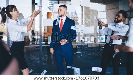 Cheerful male corporate director in formal suit standing in center of room and enjoying monetary rain with happy employees holding dollars in hand and tossing up, concept of wealthy and abundance