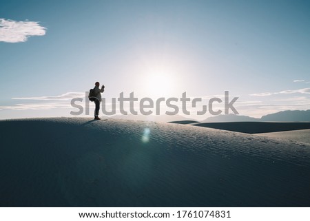 Full body of faceless male traveler in casual clothes standing on sandy dune and taking photo of amazing rest in desert in White Sands National Park
