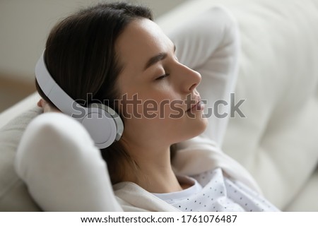 Serene young woman closed eyes relaxing leaned on couch puts hands behind head listens meditative music through modern wireless headphones, close up view. Free time, no stress, lounge at home concept Royalty-Free Stock Photo #1761076487