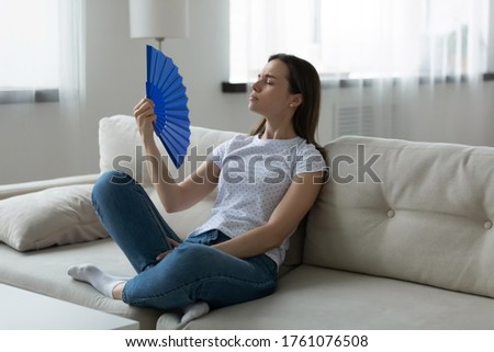Woman wear casual clothes sitting resting on couch in living room suffers from unbearable hot summer day weather waving hand blue fan reduces heat cooling herself. Home without air conditioner concept Royalty-Free Stock Photo #1761076508