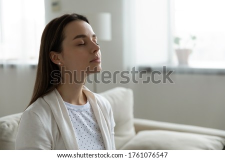 Calm serene woman resting sitting on couch in modern living room closed her eyes breath fresh humidified air. Fatigue relief repose, boost inner balance and mindfulness, meditation practice concept Royalty-Free Stock Photo #1761076547