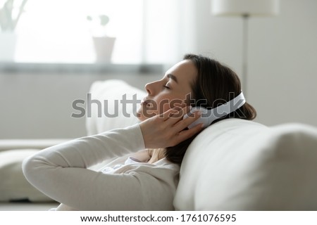 Calm female resting leaned on comfy couch in living room spend free time at home closed eyes enjoy music listening favourite song through wireless modern headphones. Pastime weekend relaxation concept Royalty-Free Stock Photo #1761076595