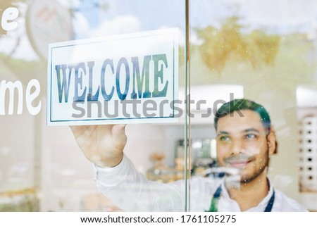 Smiling bakery shop owner sticking welcome sign on glass wall