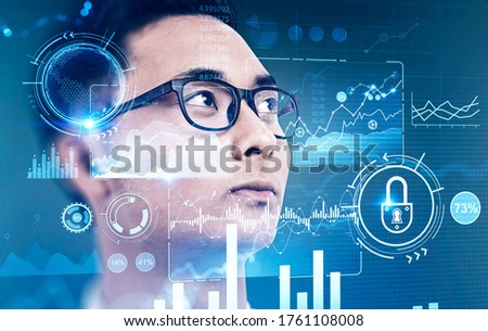 Portrait of serious young Asian businessman in glasses over blurry blue background with double exposure of immersive HUD business interface. Concept of big data and hi tech. Toned image