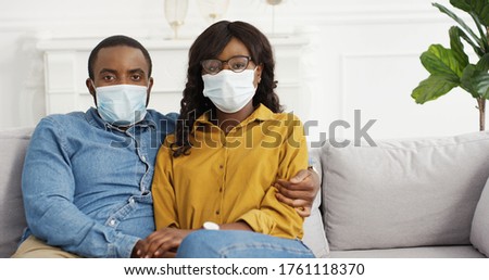 Portrait African American Couple Sitting On Sofa In Protective Medical Masks. 