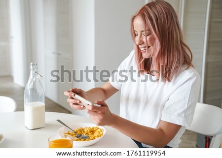 Photo of young excited woman playing online game on cellphone while having breakfast in white kitchen