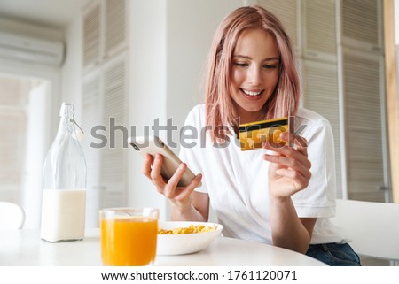 Photo of young smiling woman holding credit card and cellphone while having breakfast in white kitchen