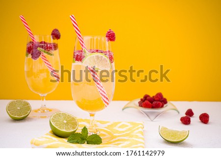 A refreshing drink with lime and raspberries in beautiful glasses stands on a white table against the background of an orange wall. On the table are slices of lime, raspberries and a bowl with berry.